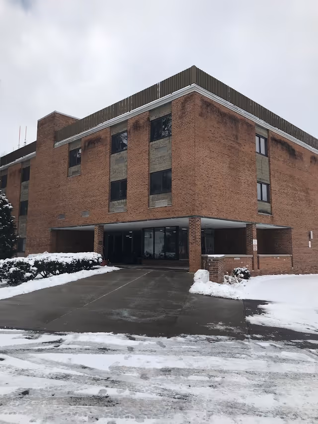 Exterior view of a three-story brick building with large windows and a covered entrance. Snow covers the ground and bushes around the building, and the sky is overcast.