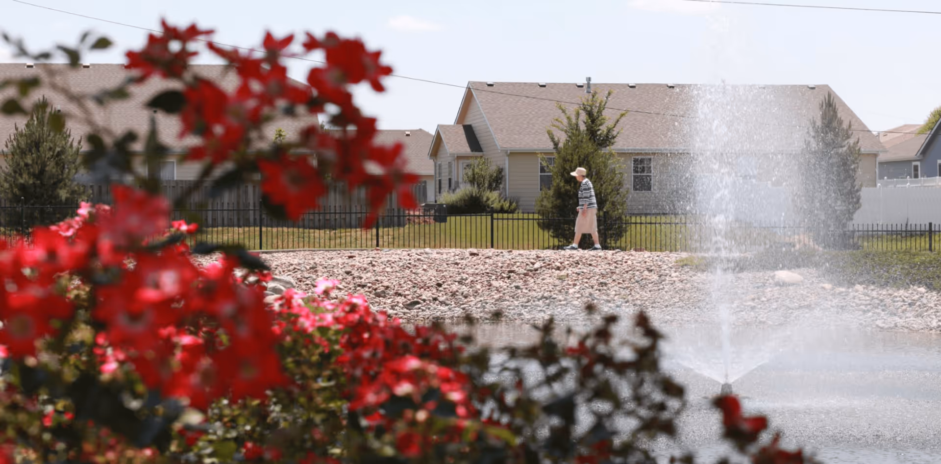 A senior woman wearing a hat and striped shirt walks along a path near a pond with a water fountain. Red flowers are in the foreground, and houses with beige siding and brown roofs are visible in the background.