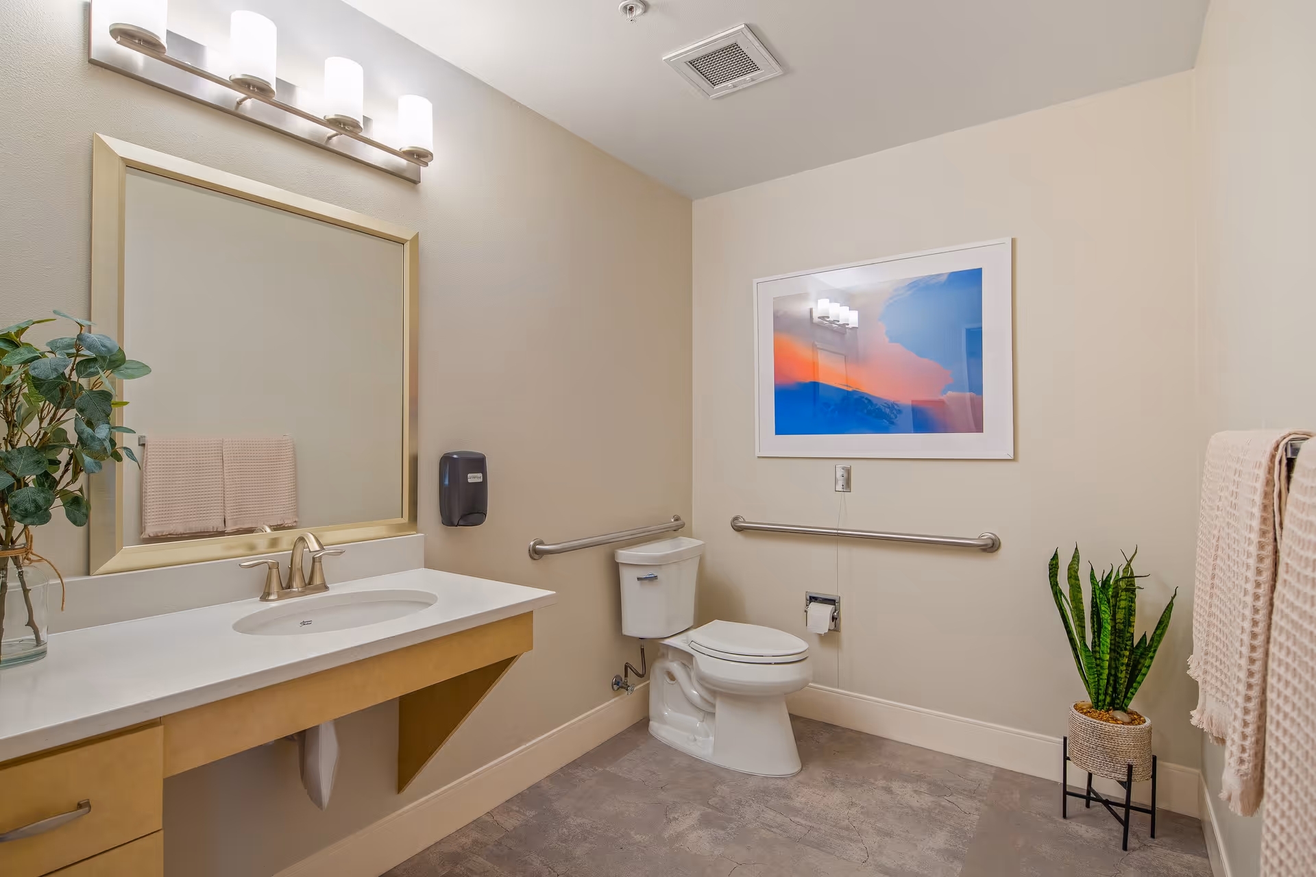 A clean and accessible bathroom with a white toilet, a large mirror above a white sink with a beige countertop, beige walls, a towel rack with beige towels, a potted plant on the floor, and a colorful framed picture on the wall.