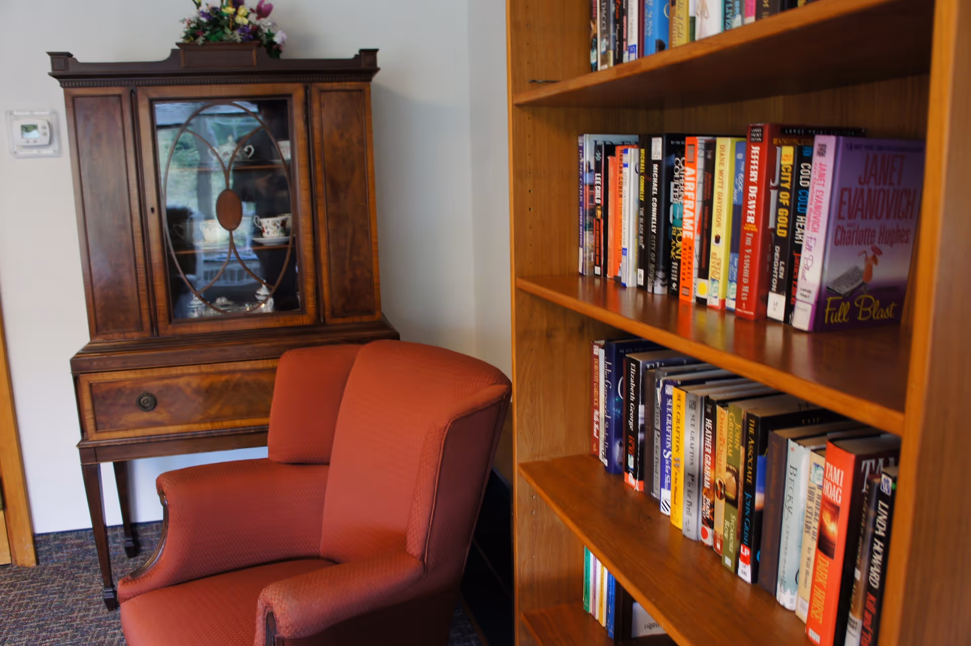 A cozy corner of a room featuring a red upholstered armchair, a wooden bookshelf filled with various books, and a wooden cabinet with glass doors displaying teacups and saucers. The room has a carpeted floor and light-colored walls.