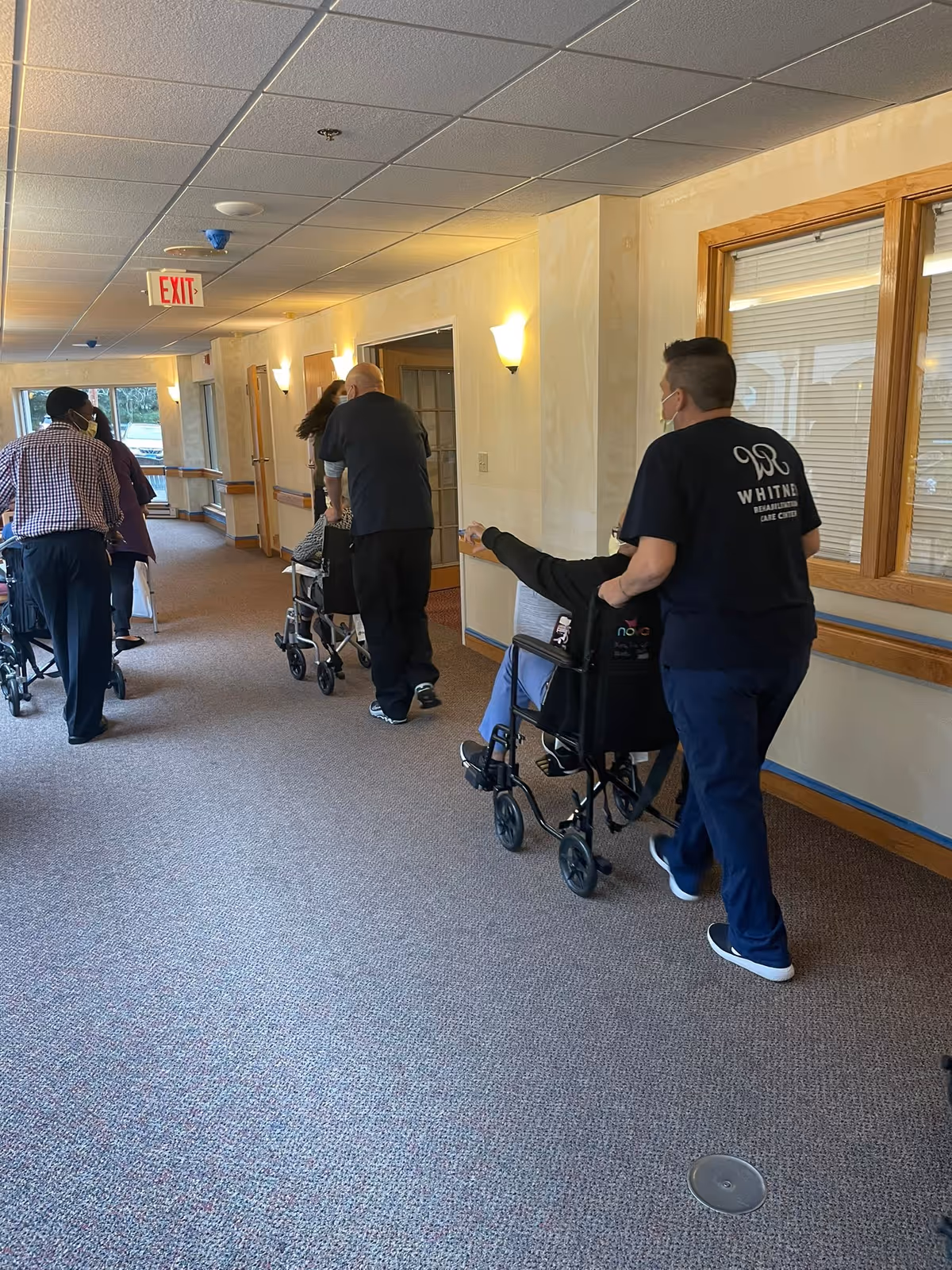 A hallway in a care facility with several people, some using walkers and one person in a wheelchair being pushed by a staff member wearing a Whitney Rehabilitation Care Center shirt. The hallway has beige walls, carpeted floor, wall-mounted lights, and windows on the right side.