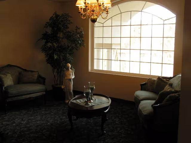 A dimly lit living room area with two upholstered sofas facing each other, a round wooden coffee table with decorative items on it, a large arched window letting in natural light, a chandelier hanging from the ceiling, and a potted plant with a small statue beside it.