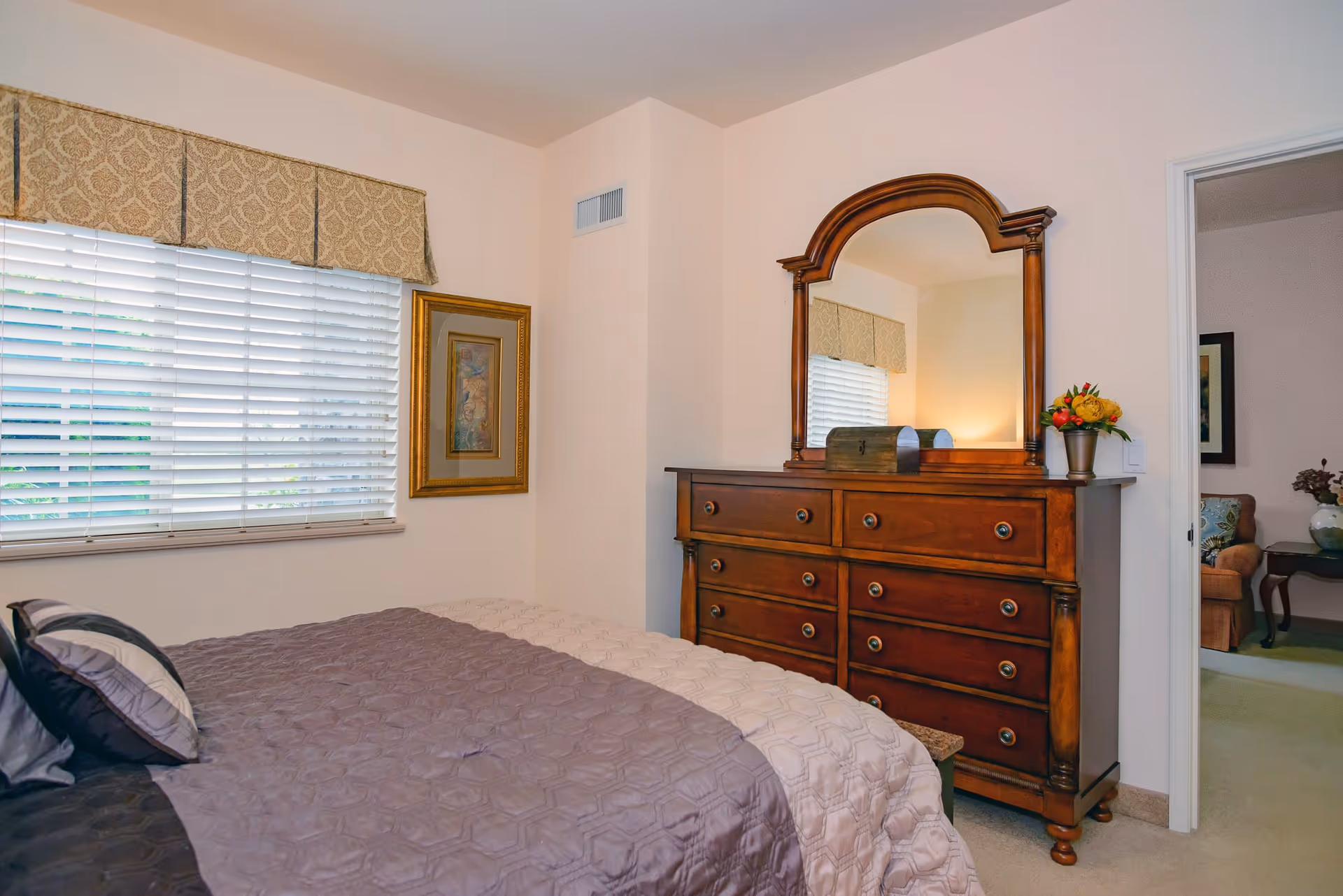 A bedroom with a bed covered in a quilted bedspread in shades of gray and beige, a wooden dresser with a large mirror on top, a small decorative chest and a vase with flowers on the dresser. There is a window with white blinds and a patterned valance, and a framed picture on the wall. An open door reveals a glimpse of a living room with a sofa and a side table with a vase.