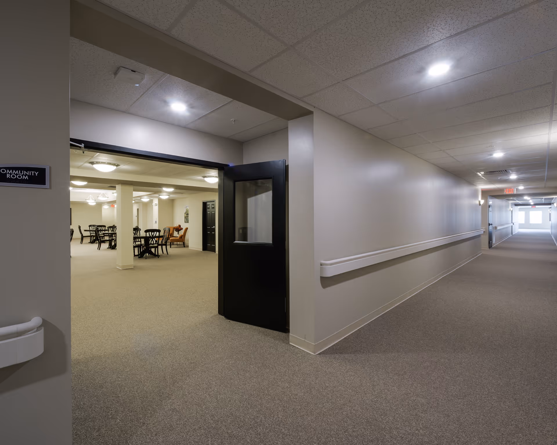 View of a hallway in Morton Estates leading to an open door labeled 'Community Room'. Inside the community room, there are several tables and chairs arranged for seating. The hallway and room have beige walls and carpeted floors, with ceiling lights providing illumination.