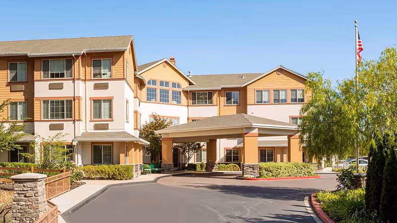 Exterior view of a multi-story senior living facility building with beige and light brown siding, large windows, a covered entrance, landscaped bushes, trees, and an American flag on a flagpole under a clear blue sky.