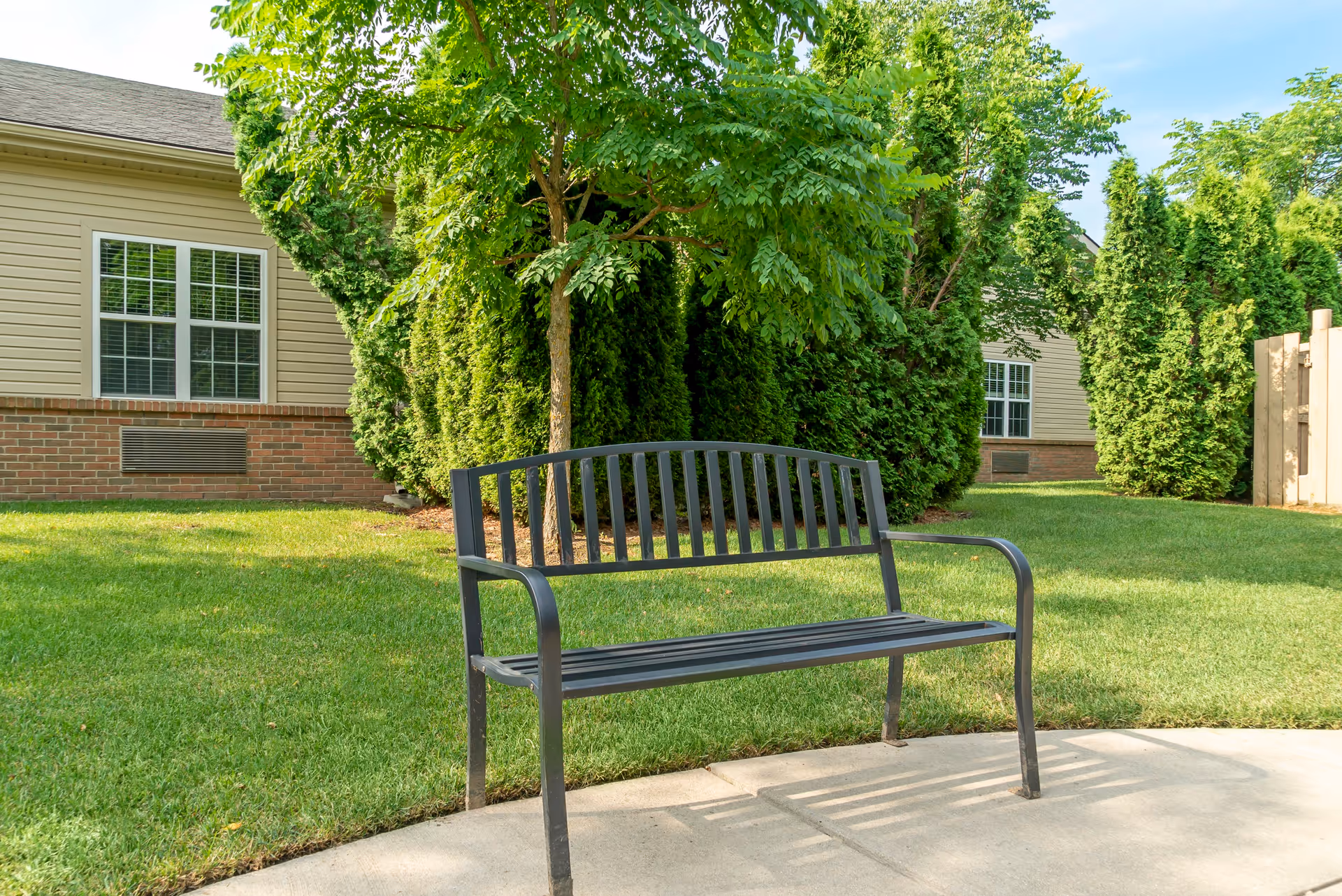 A black metal bench on a concrete path in a grassy outdoor area with trees and shrubs, adjacent to a beige building with white-framed windows.