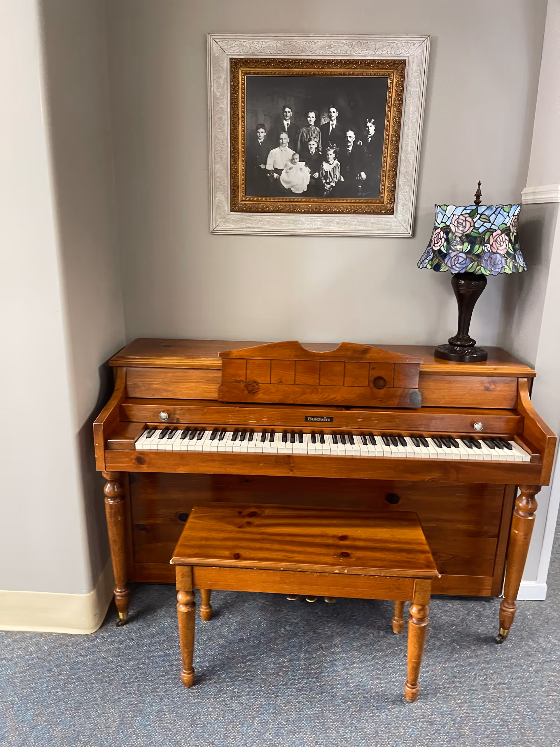 A wooden upright piano with a matching bench in front of it, placed against a light gray wall. Above the piano hangs a framed black and white family portrait. To the right on top of the piano is a decorative lamp with a stained glass floral lampshade.