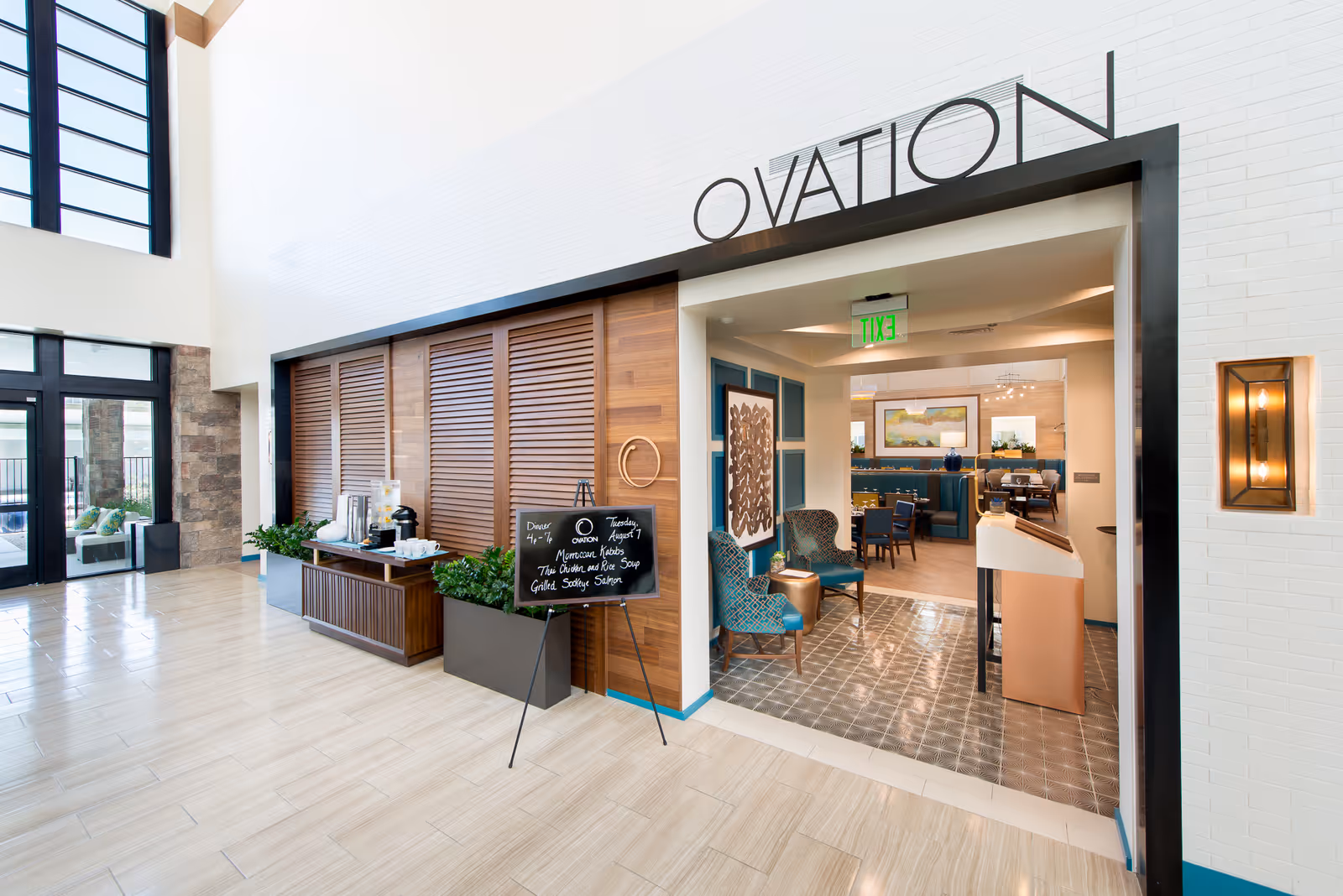 Entrance to a dining area named Ovation in a senior living facility. The entrance has a black frame with the word OVATION above it. Inside, there are tables and chairs arranged for dining, with artwork on the walls and a cozy atmosphere. Outside the entrance, there is a small table with coffee and cups, and a chalkboard sign listing the dinner menu. The surrounding area has large windows and light-colored tiled flooring.