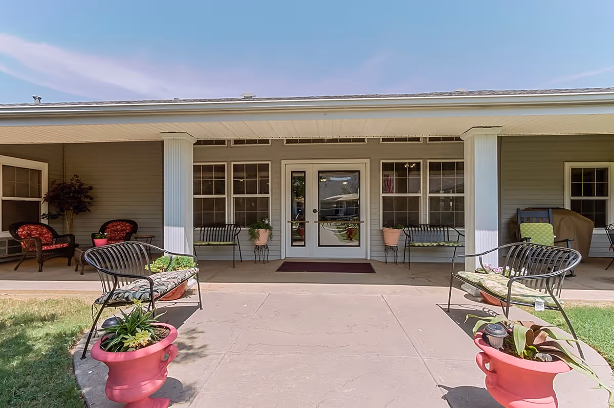 Covered front entrance of a senior living facility with patio seating, planters, and a central glass door.