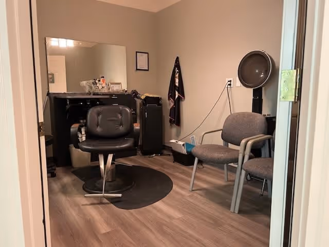 Interior view of a small salon or hair styling room with a black salon chair on a black mat, a large mirror on the wall, a hair dryer hood, two gray chairs, and various salon supplies on a counter and shelf.