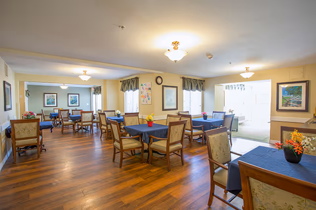 Bright communal dining room with multiple tables and chairs, blue tablecloths, hardwood floors, and framed artwork on the walls.