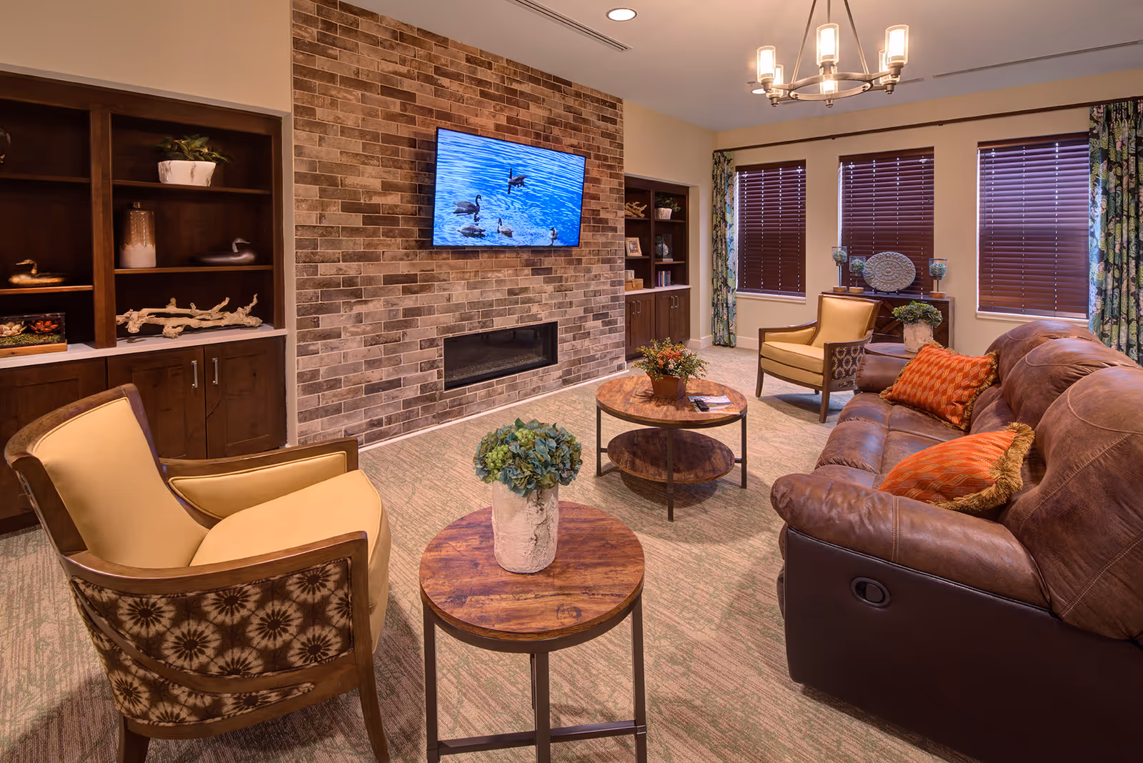 Cozy living room with a leather sofa, armchairs, round wooden coffee tables, a brick fireplace with a wall-mounted TV, built-in shelving, and windows with blinds.