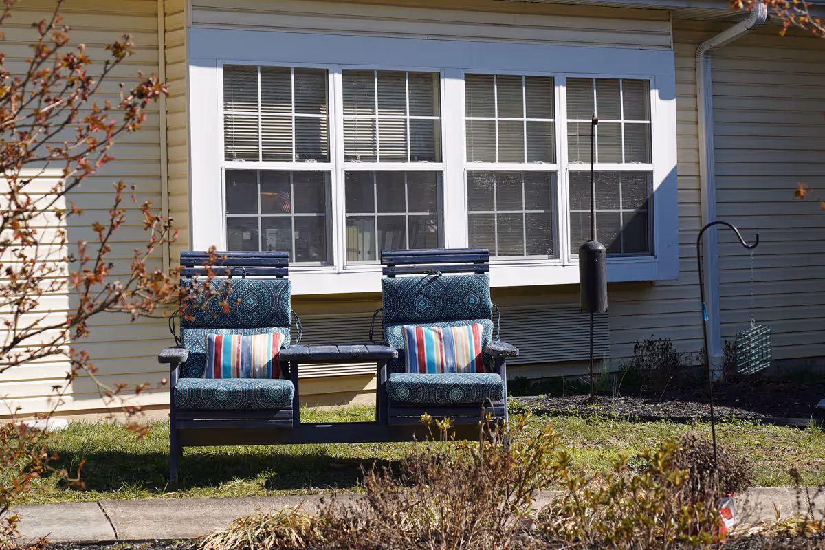 Outdoor seating area with two cushioned chairs connected by a small table, placed on grass in front of a building with beige siding and large windows. There are decorative pillows on the chairs and a bird feeder hanging nearby.