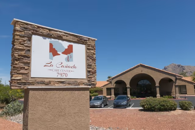 Exterior view of La Cañada Care Center building with a stone and stucco sign displaying the facility name and number 7970. The building has a tiled roof and arched entrance, with a parking lot and two cars visible in front. Mountains and clear blue sky are in the background.