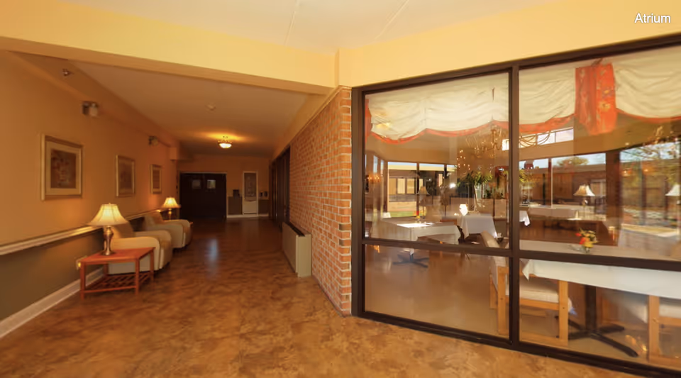 Interior view of a hallway in a senior living facility with beige walls, framed artwork, and two armchairs with side tables and lamps. To the right, large glass windows reveal a dining area with tables covered in white tablecloths, chairs, and floral centerpieces. The floor has a brown patterned surface, and the ceiling has recessed lighting.