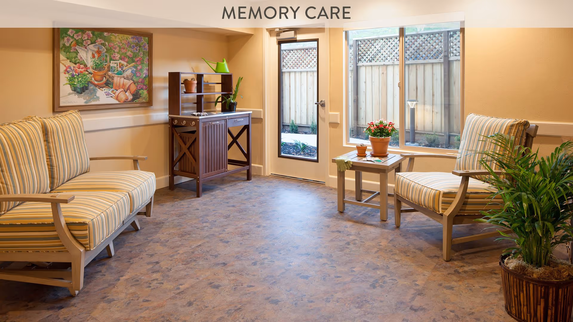 A cozy memory care room with two striped cushioned armchairs and a small table with potted plants. There is a wooden cabinet with shelves holding more plants and decor. A large window and glass door provide natural light and a view of a fenced outdoor area. A colorful painting hangs on the wall.