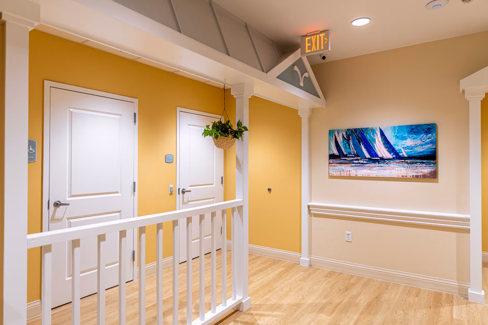 Interior hallway in a senior living facility with yellow and beige walls, two white doors, a hanging plant, a colorful painting of sailboats on the wall, wooden flooring, and an illuminated exit sign above.