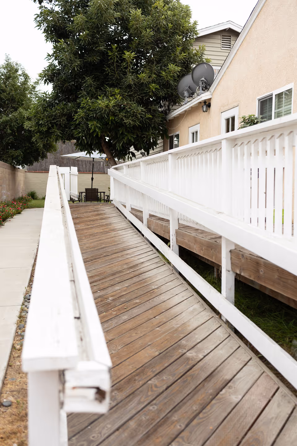 A wooden wheelchair ramp with white railings leading to the entrance of a beige building with windows and satellite dishes, surrounded by greenery including a large tree and some plants along a concrete pathway.