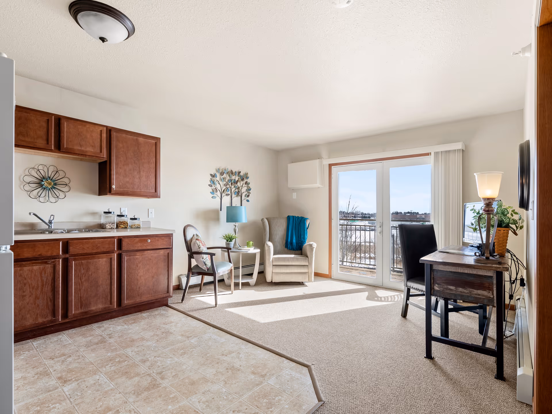 A bright senior living room with a small kitchenette on the left featuring wooden cabinets and a sink. The room has a beige carpeted area with a comfortable armchair, a wooden chair, a small side table with a lamp, and a desk with a chair and a lamp on the right. Large glass doors open to a balcony with a view outside.