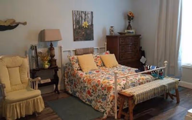 Cozy bedroom with a floral bedspread and yellow pillows, armchair, wooden dresser, and bench at the foot of the bed.