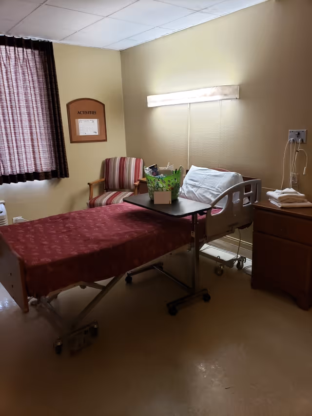 A small room with a hospital bed covered with a red patterned blanket, a pillow, and an adjustable overbed table holding a green basket with items. There is a striped armchair next to the bed, a wooden nightstand with folded towels, and a wall-mounted light above the bed. A window with dark curtains and an 'Activities' bulletin board are visible on the wall.