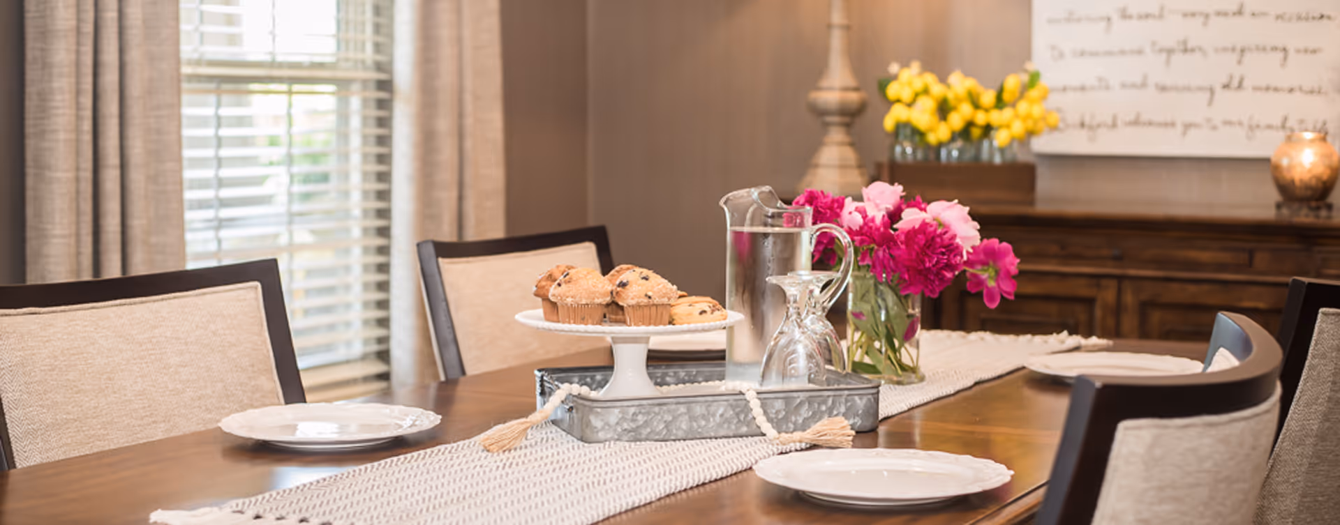 Dining room table arranged with plates, a pitcher, a tiered plate of muffins, and vases of flowers in a warmly lit room.