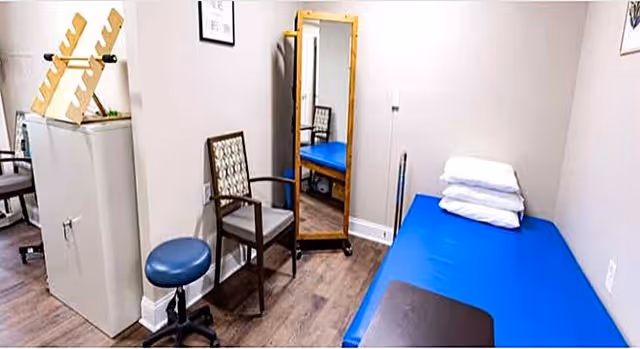 A small room with a blue examination table covered with folded white towels, a wooden chair with a patterned cushion, a blue round stool on wheels, a tall standing mirror, and a small cabinet with a wooden rack on top. The room has light-colored walls and wood flooring.