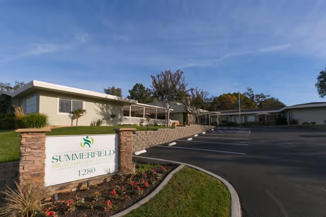 Entrance and parking lot of Summerfield Healthcare Center with its sign in front of a single-story building under a blue sky.