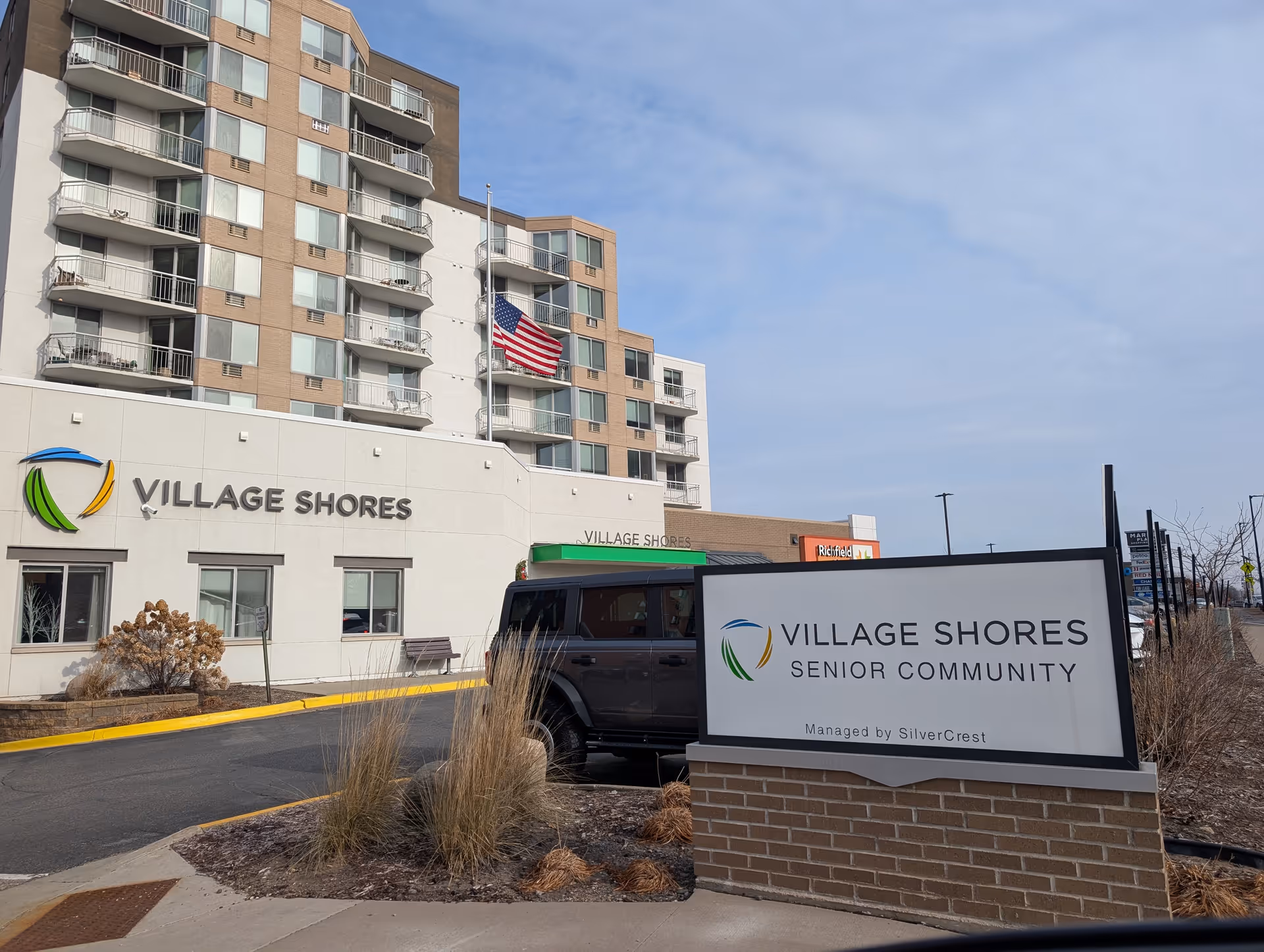 Exterior view of Village Shores Senior Community building with multiple floors and balconies. An American flag is flying at half-mast in front of the building. A large sign in the foreground reads 'Village Shores Senior Community Managed by SilverCrest'. There is a black vehicle parked near the entrance and some dry landscaping with bushes and grasses.