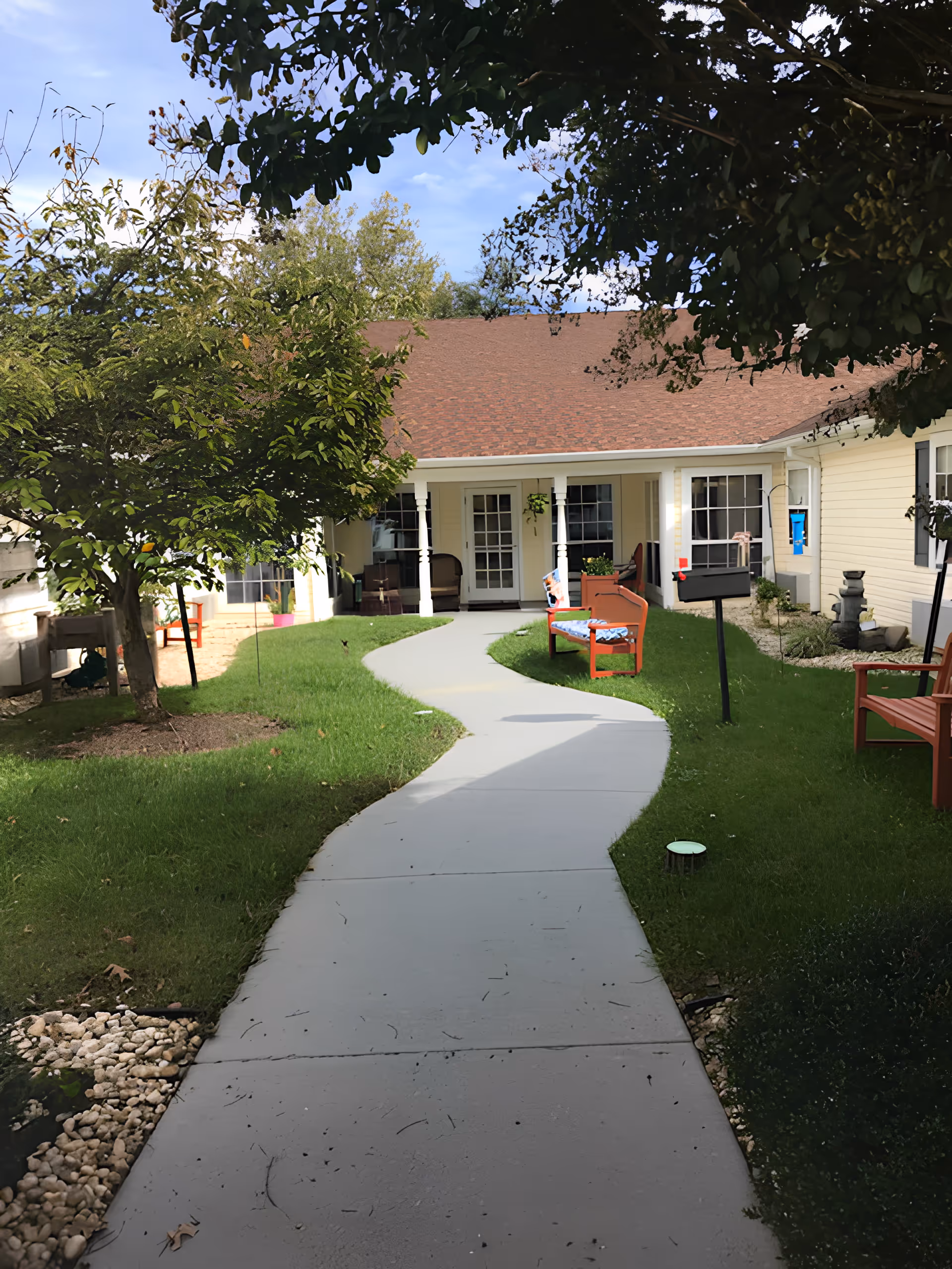A curved concrete pathway leads through a green lawn with trees and red benches towards a single-story building with a brown shingled roof and cream-colored siding. The building has multiple windows and a door with glass panes. The area is shaded by tree branches overhead.