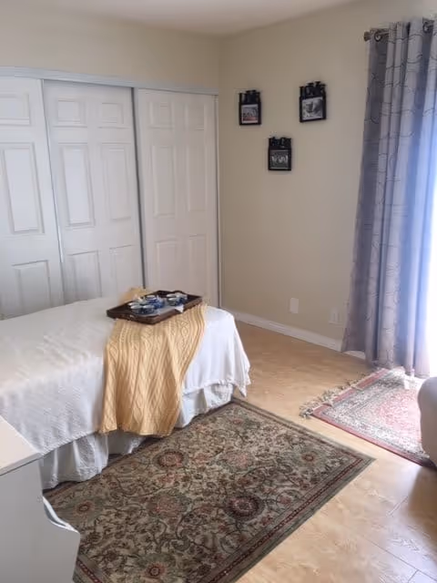 A bedroom with a bed covered in white bedding and a yellow throw blanket. A tray with cups and a teapot is placed on the bed. The room has light-colored walls, a set of white sliding closet doors, three small framed pictures on the wall, a patterned area rug on the wooden floor, and a window with gray curtains allowing natural light to enter.