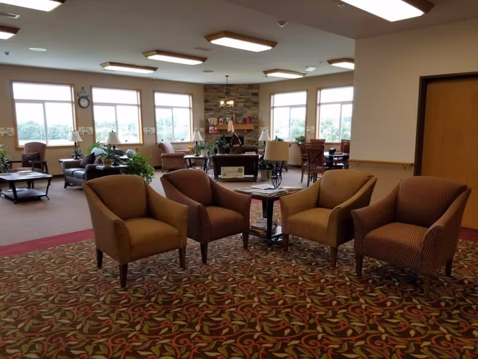 A spacious senior living common area with multiple armchairs arranged around small tables on a patterned carpet. Large windows let in natural light, and there is a stone fireplace in the background with decorative items on the mantle. Several lamps and plants are placed throughout the room, creating a cozy and inviting atmosphere.