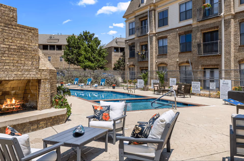 Outdoor pool area at Discovery Village At Castle Hills featuring a swimming pool surrounded by lounge chairs with blue cushions, a brick fireplace with a fire burning, and several cushioned chairs with floral pillows arranged around a gray table. The background shows a multi-story brick building with balconies and windows, and a clear blue sky with a few clouds.