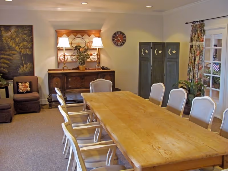 A well-lit dining room with a long wooden table surrounded by white cushioned chairs. Against the far wall is a wooden sideboard with two lamps, a plant, and decorative plates. A clock hangs on the wall above a decorative folding screen with moon and star cutouts. To the right, floral curtains frame glass doors leading to another room.