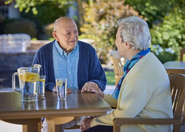 An elderly man and woman sitting at an outdoor table, holding hands and smiling at each other. The table has a pitcher and glasses of water with lemon slices. The background shows greenery and sunlight.