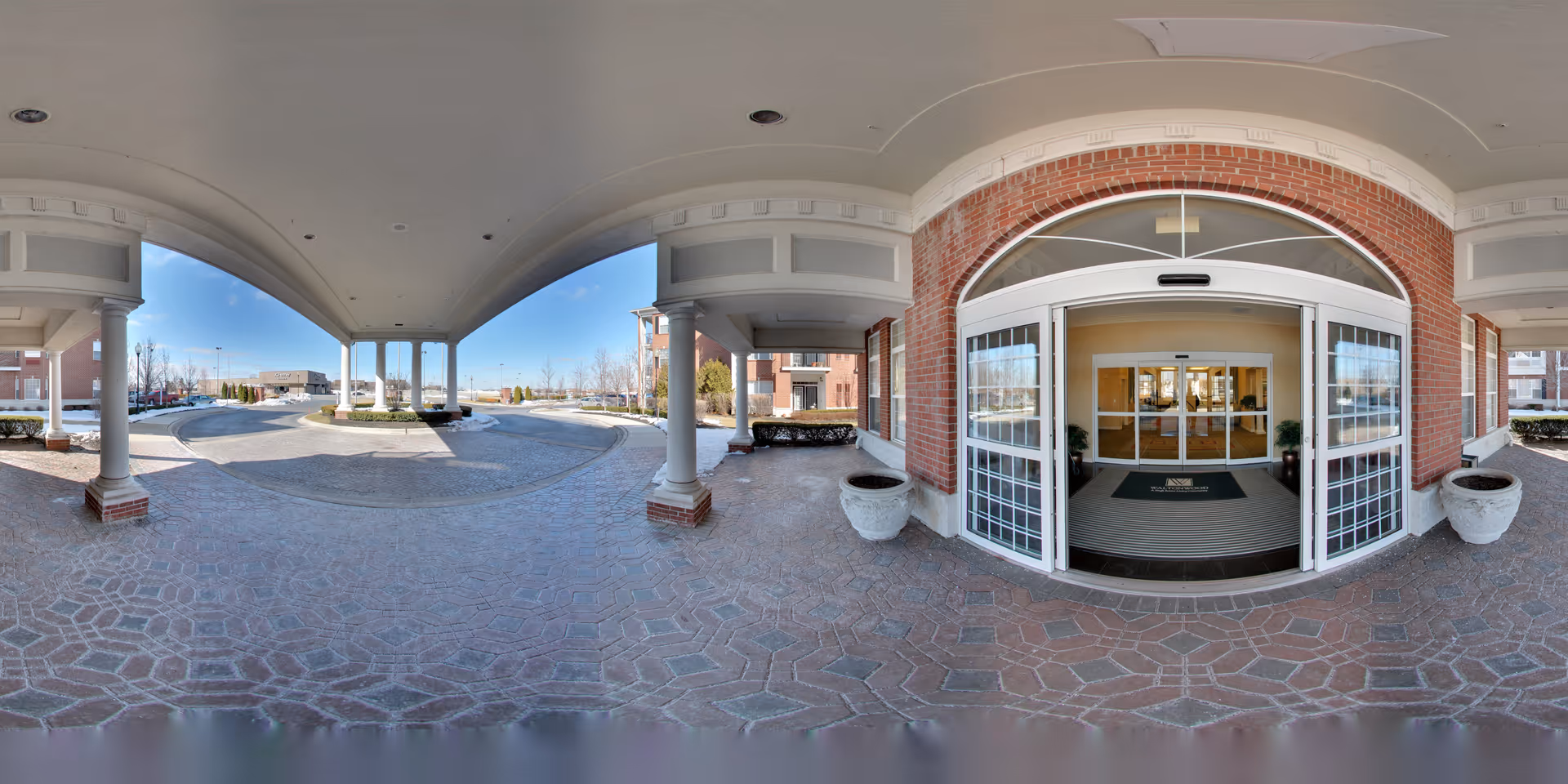 Covered entrance area of Waltonwood Lakeside facility with brick walls and white columns. Automatic sliding glass doors lead into the building, and there are large planters on either side of the entrance. The driveway curves around the entrance with parking and landscaping visible in the background under a clear blue sky.