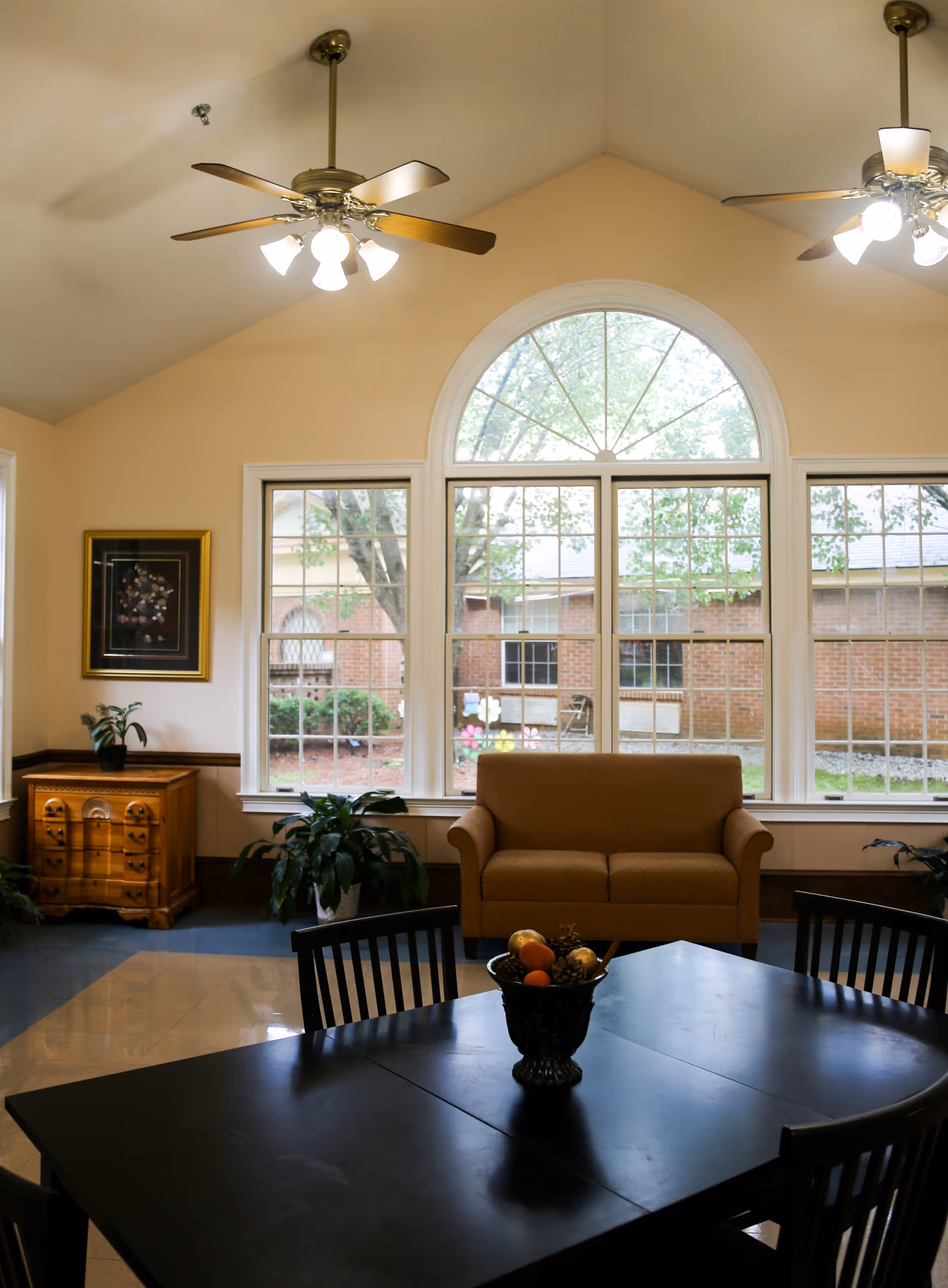 A cozy interior room with a large arched window showing an outdoor view of trees and a brick building. The room features a brown loveseat, a black dining table with chairs, a wooden cabinet with a plant on top, two ceiling fans with lights, and several potted plants.