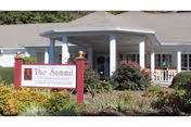 Exterior view of The Summit of Lawrenceburg senior living facility showing the entrance with a covered porch, surrounded by landscaped bushes and flowers, and a sign in front displaying the facility's name.