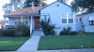 Single-story residential building with a front porch, a red door, and several windows. The house is surrounded by a well-maintained lawn with a concrete walkway leading to the front steps. There are bushes and plants near the house, and another building is partially visible on the right side.