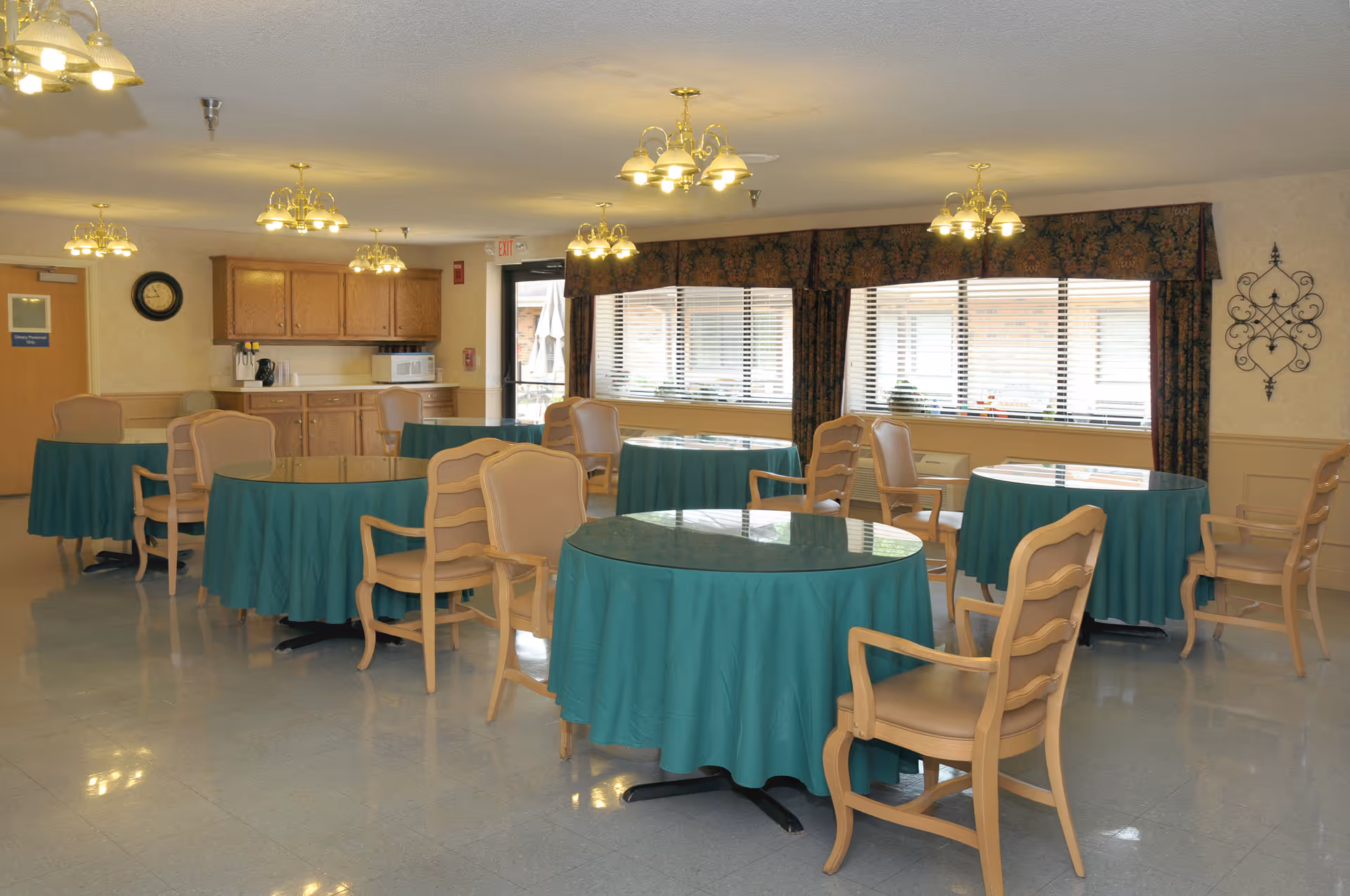 A dining room with several round tables covered with green tablecloths and surrounded by beige chairs. The room has multiple ceiling light fixtures, large windows with blinds and patterned valances, wooden cabinets, a microwave, and a clock on the wall.