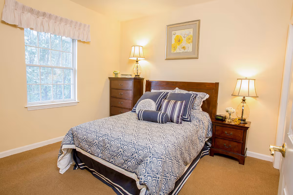 A cozy bedroom with a double bed featuring blue and white patterned bedding and multiple pillows. There is a wooden headboard, a wooden nightstand with a lamp, a small vase with flowers, and an alarm clock. A wooden chest of drawers with a lamp and small plant is positioned against the wall. A window with a white valance lets in natural light, and a framed floral artwork hangs above the bed.