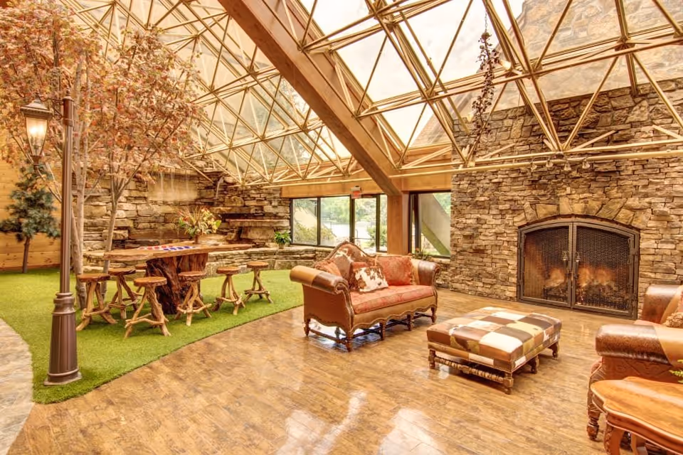 A cozy indoor living space with a glass ceiling and stone walls. The room features a stone fireplace with a fire burning, a brown leather sofa with patterned cushions, a matching ottoman, and a rustic wooden table with stools on a patch of artificial grass. There are also decorative trees and a vintage-style street lamp inside the room.