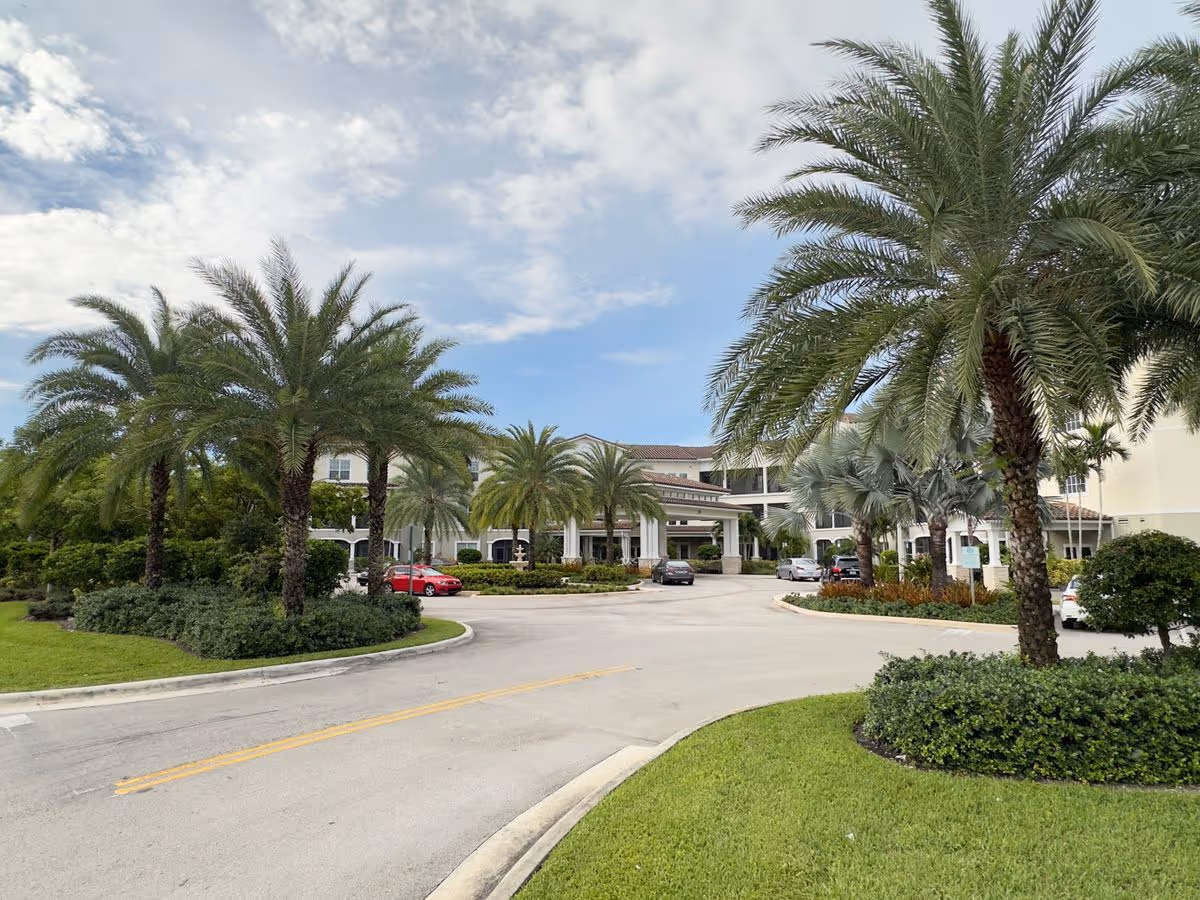 Front entrance of a senior living facility with palm trees, a circular driveway and parked cars under a partly cloudy sky.