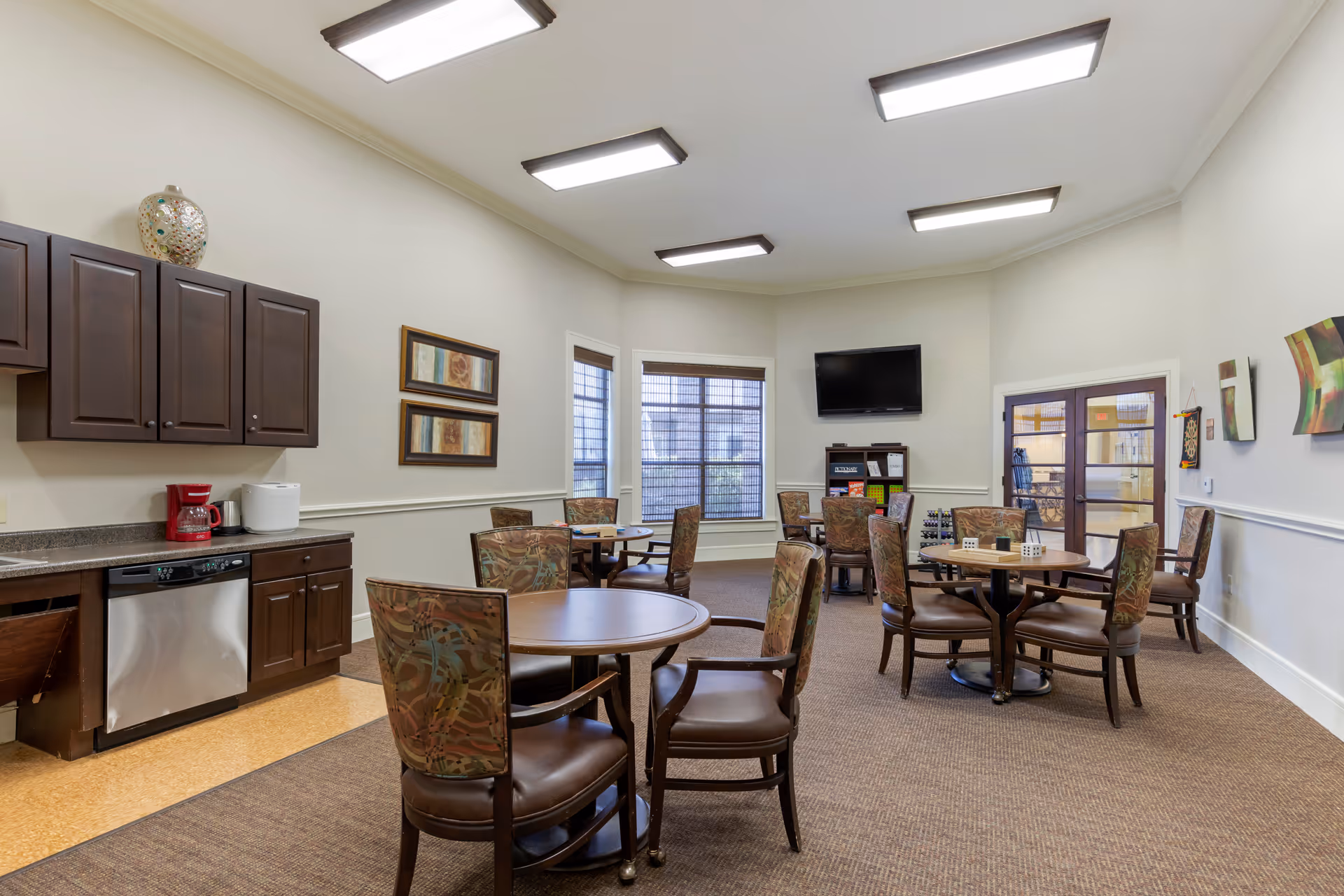 A well-lit dining room in an assisted living facility with round wooden tables and cushioned chairs arranged neatly. The room features a small kitchenette area with dark wood cabinets, a coffee maker, and a dishwasher. There are framed artworks on the walls, a flat-screen TV mounted on the far wall, and large windows letting in natural light.
