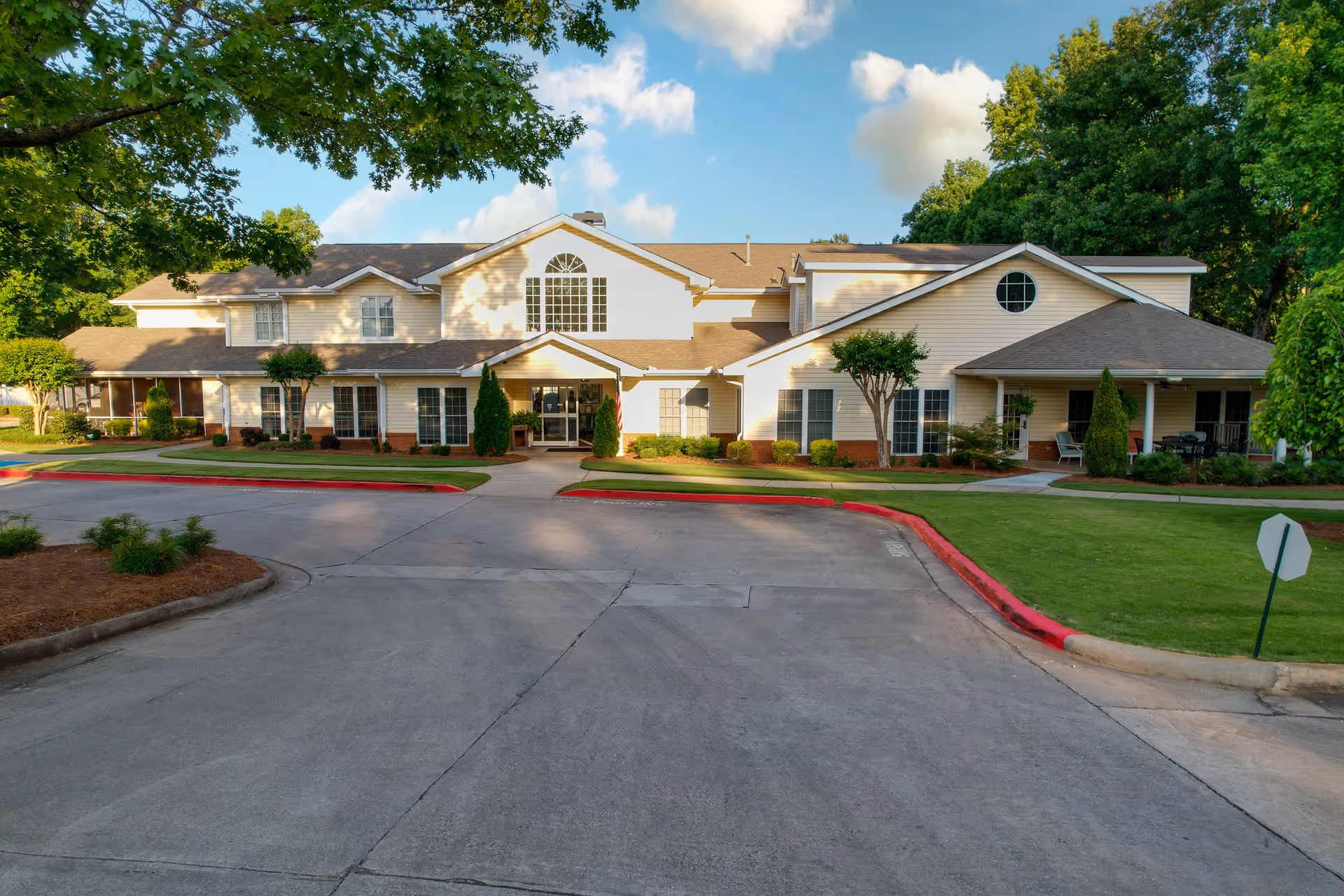 Front exterior view of a two-story senior living facility building with beige siding, multiple windows, a central entrance with a small covered porch, surrounded by green lawns, trees, and a paved driveway with red curbs.