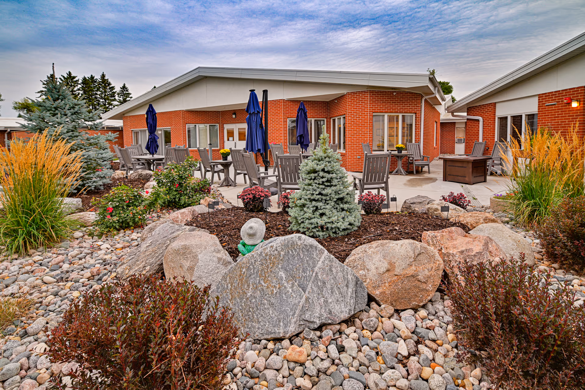 Outdoor patio area at a senior living facility featuring multiple tables and chairs with closed blue umbrellas. The patio is surrounded by landscaped garden beds with rocks, shrubs, small trees, and ornamental grasses. The building has red brick walls and white trim under a partly cloudy sky.