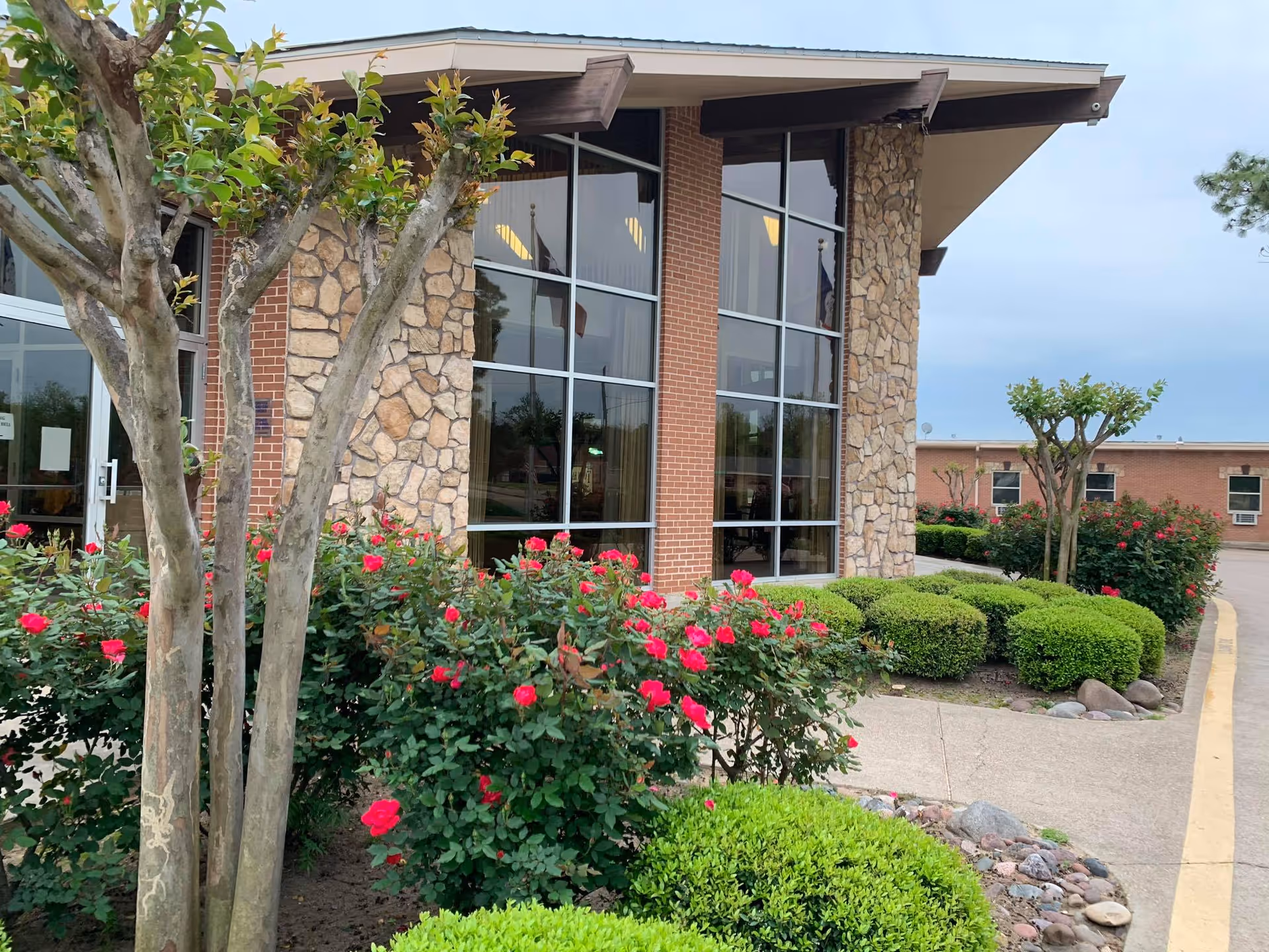Exterior view of a building with large windows, stone and brick walls, surrounded by neatly trimmed bushes and blooming red flowers under a cloudy sky.