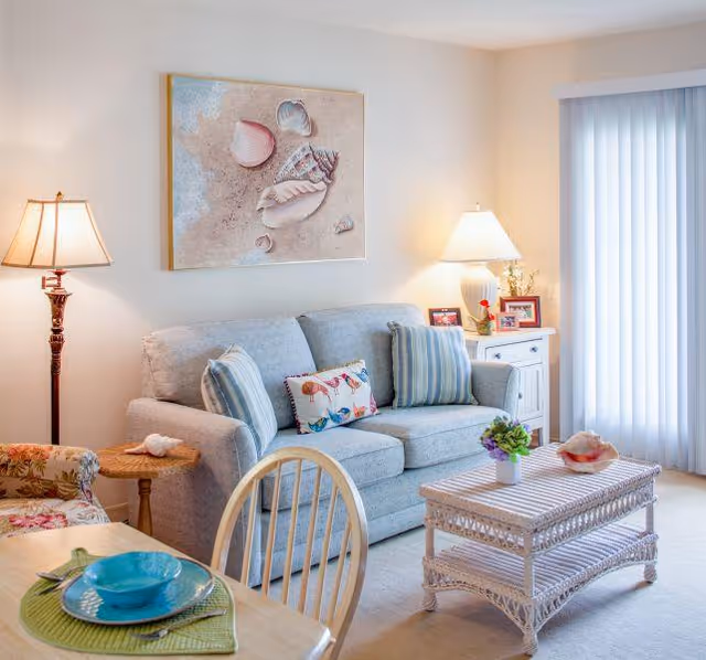A cozy living room with a light gray sofa adorned with striped and patterned pillows, a wicker coffee table with a small plant and decorative shell, a floral armchair, a wooden dining table set with turquoise dishes, two table lamps, a side table with framed photos, and a large painting of seashells on the wall. Vertical blinds cover a sliding glass door.