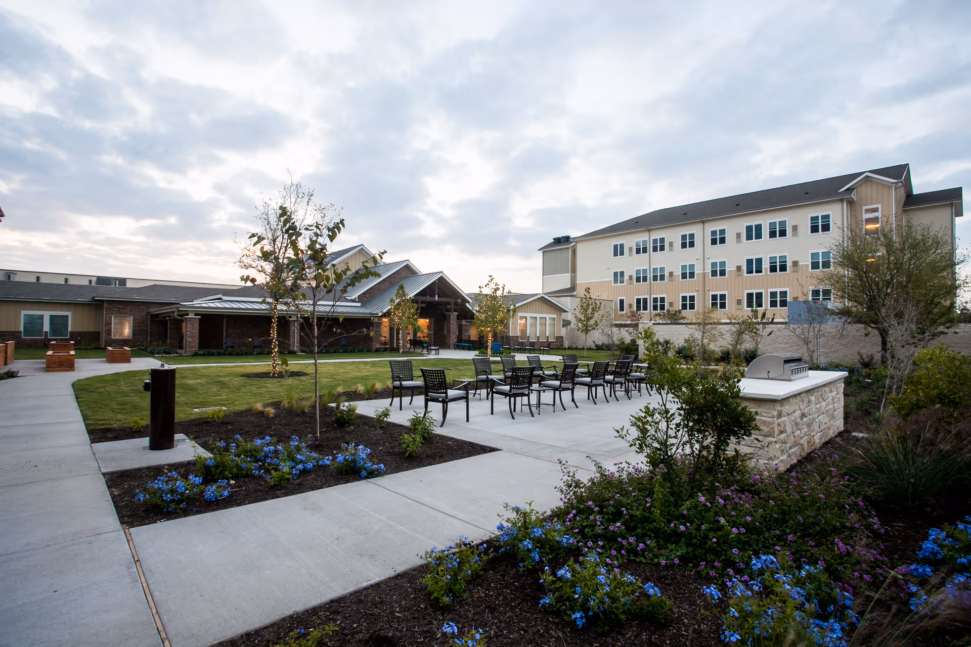 Outdoor courtyard with patio tables and chairs, landscaped flower beds, and a multi-story senior living building in the background.