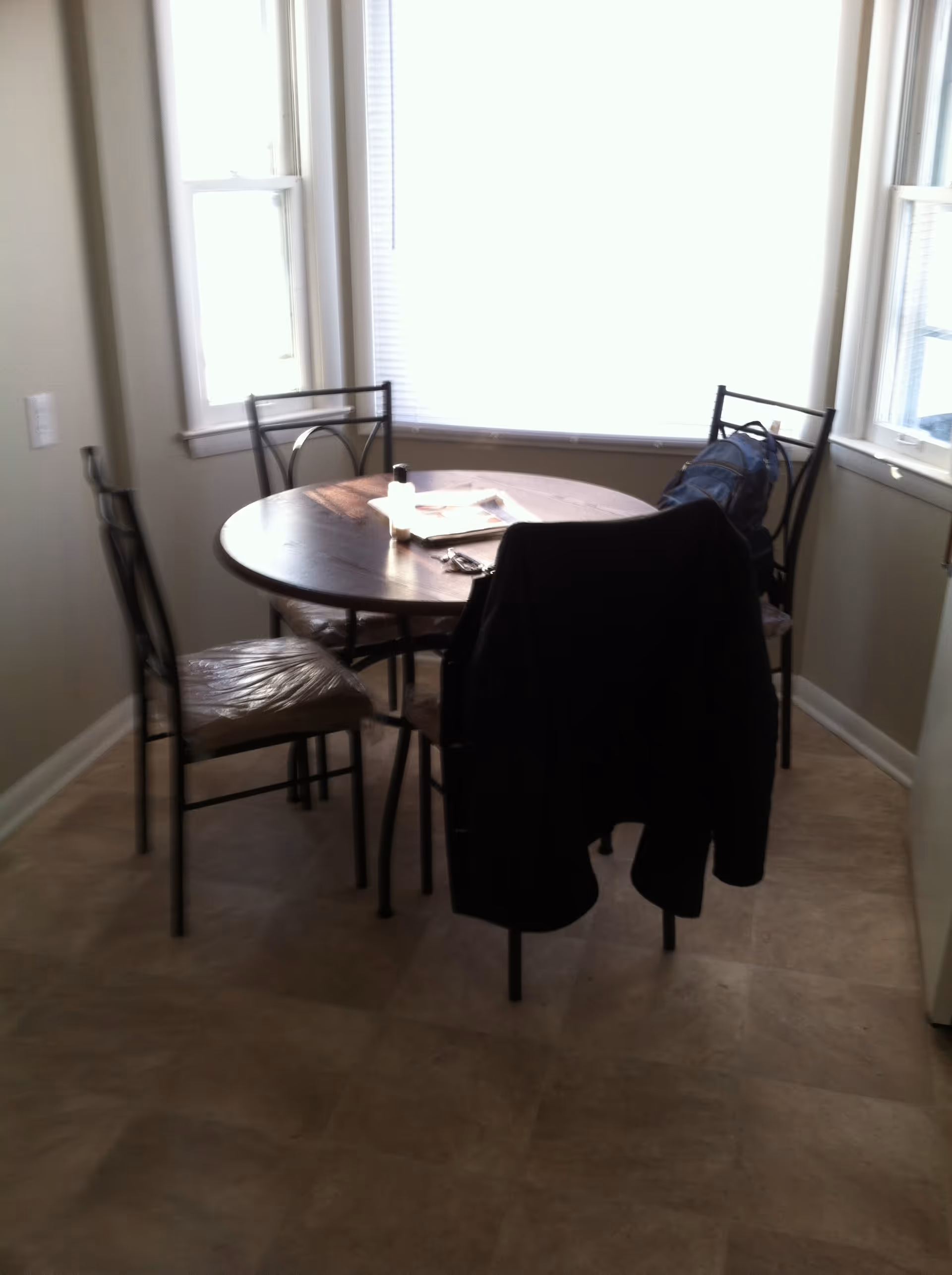 A small dining area with a round wooden table and four metal chairs with padded seats. One chair has a black jacket draped over it, and another has a blue backpack. The table has some items on it, including a newspaper and a small bottle. The room has beige walls and three windows with white blinds, letting in bright natural light.